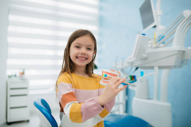 Child smiling in a dental clinic, holding a toothbrush and teeth model while seated in a dental chair.